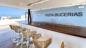A bar area with wicker chairs and a wooden counter, a sign reading 'VISTA BUCERIAS,' and a view of the ocean. - Ocean-view Condo With Infinity Pool, Rooftop Lounge, & Private Balcony (Bucerías)