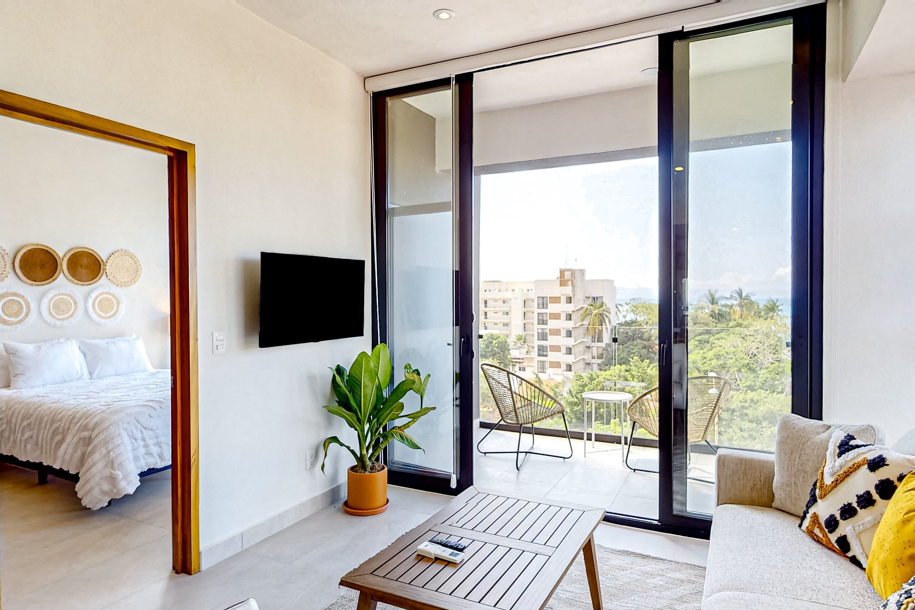 Una sala de estar moderna con sofá, una mesa de centro, una planta en maceta, televisión y un balcón con silla y mesa.