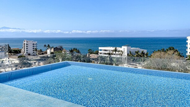 A clear blue swimming pool with a view of the ocean and buildings in the background. - Ocean-view Condo With Infinity Pool, Rooftop Lounge, & Private Balcony (Bucerías)