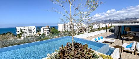 An infinity pool with a clear view of the sea, surrounded by lounge chairs and a small tree.