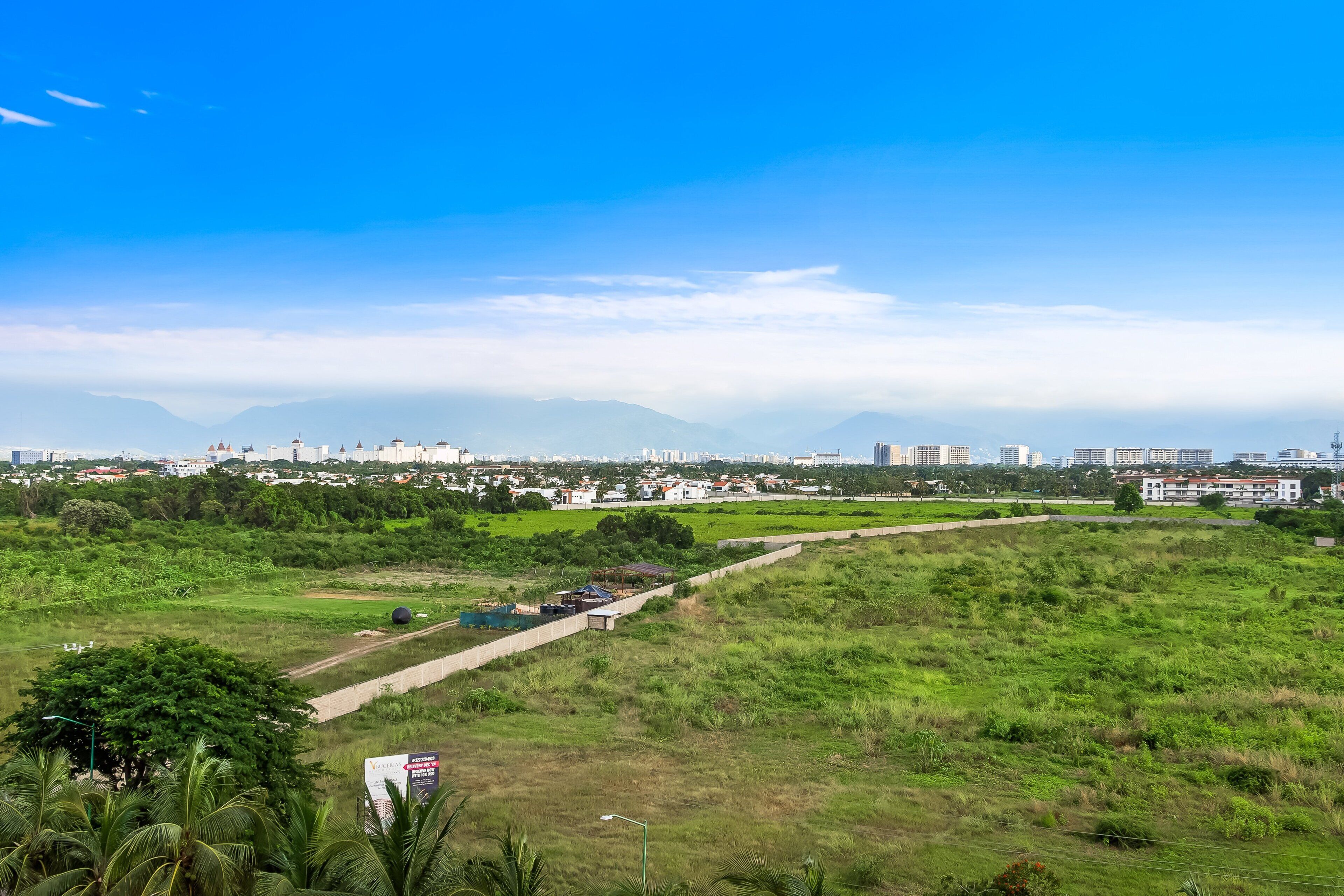 Un paisaje urbano con un gran parque verde, un camino pavimentado y montañas a la distancia.