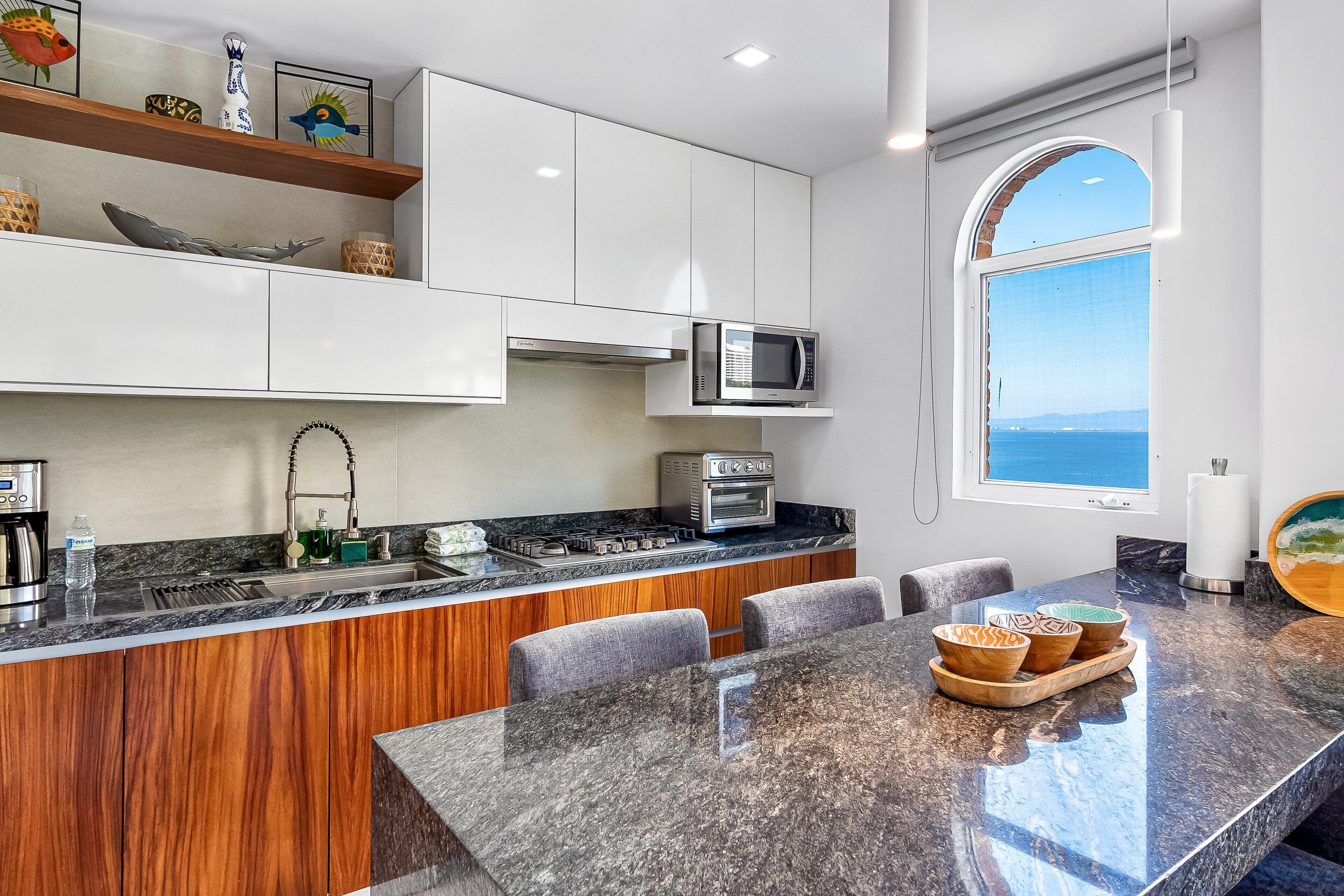 Una cucina moderna con mobili bianchi, un piano di lavoro in granito, forno integrato e vista sul mare attraverso un'ampia finestra.