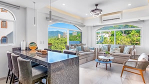 A modern living room with a large island, seating area, and large windows with a view of greenery and buildings. - Charming Beach Apartment With Pool and Beach Access, Full Kitchen and Wifi (Puerto Vallarta)