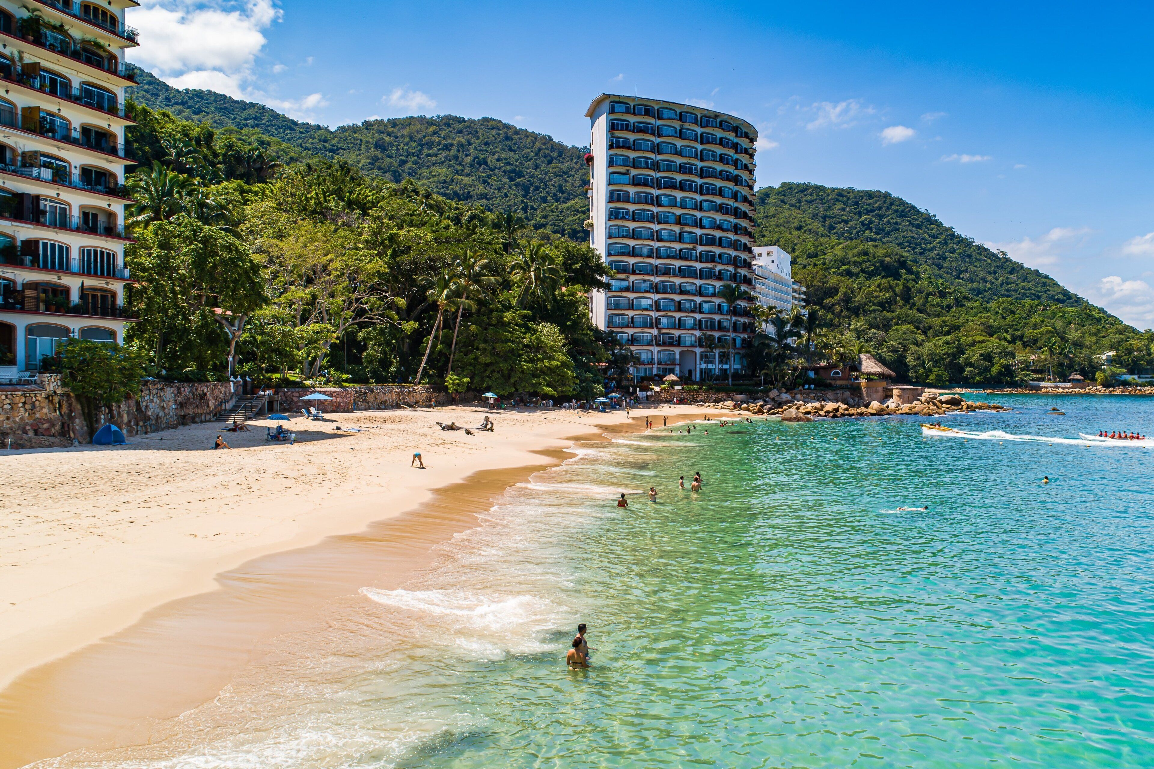 A beach with clear turquoise water, a sandy shore, and a hotel with multiple balconies overlooking the beach.