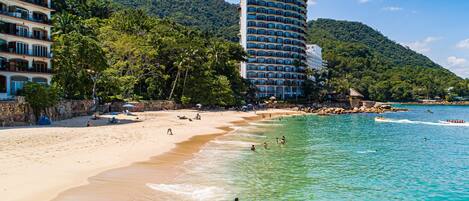 A beach with clear turquoise water, a sandy shore, and a hotel with multiple balconies overlooking the beach.