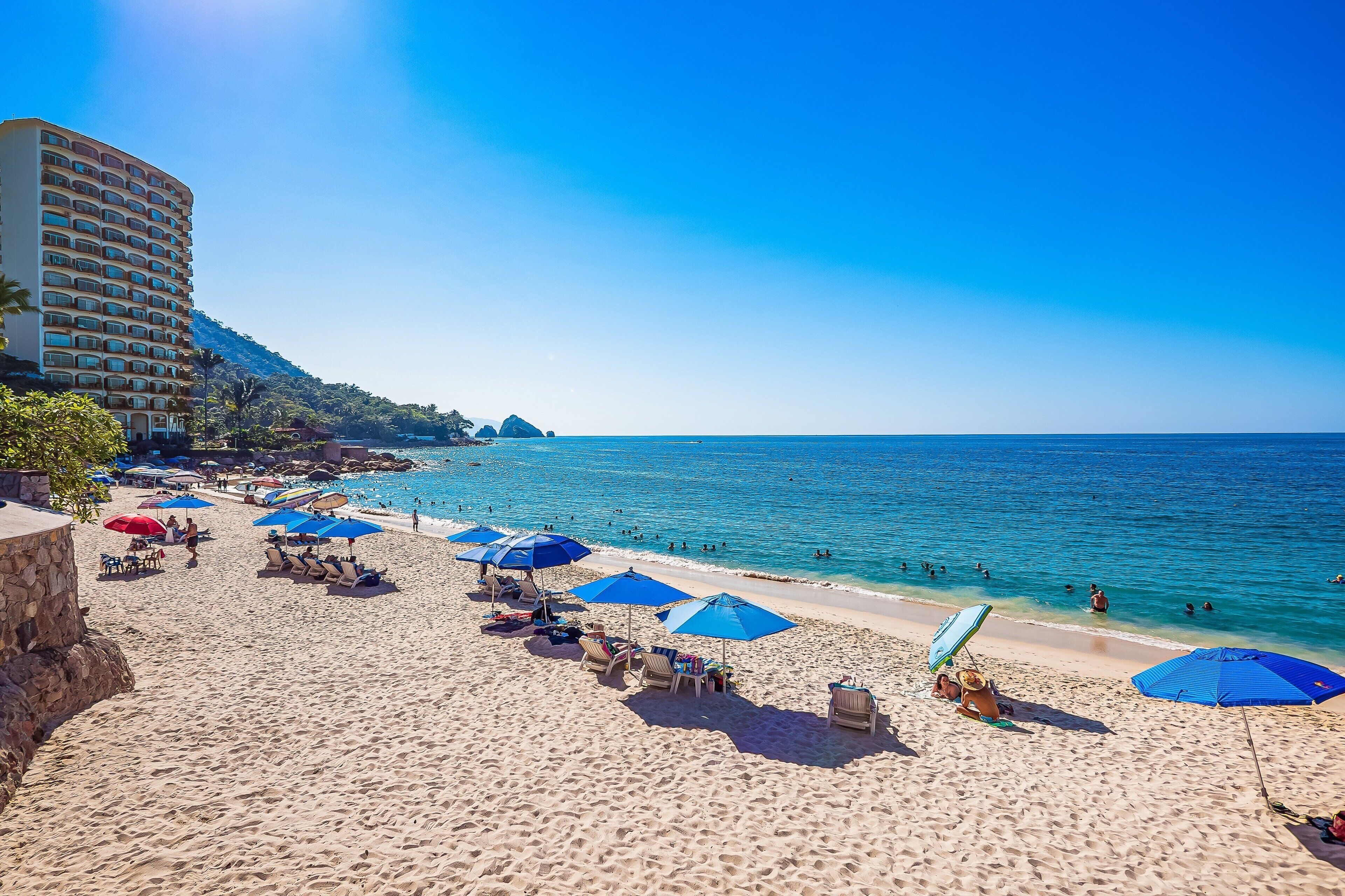 A beach with sun loungers and umbrellas, a hotel building, and clear blue water.