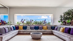 Ein modernes Interieur mit einer Betonbank, einem großen Fenster mit Blick auf Pflanzen und einer Sitzecke mit verschiedenen Kissen.