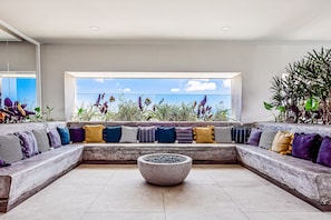 A modern interior with a concrete bench, a large window with a view of plants, and a seating area with various cushions. - Ocean-view Condo in Puerto Vallarta With Rooftop Pool, Fitness Room, and Balcony (Puerto Vallarta)