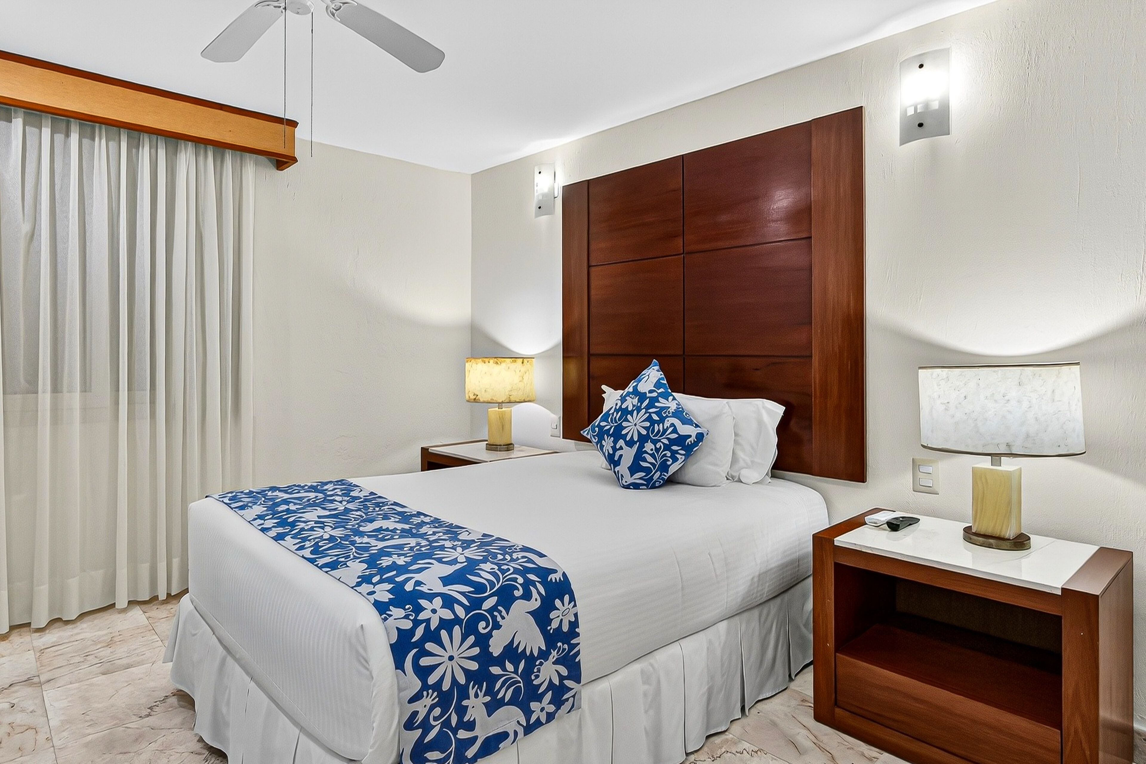 Habitación de hotel con una cama grande, cabecero de madera, dos mesitas de noche con lámparas, un ventilador de techo y una ventana con cortinas.