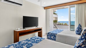 A hotel room with a flat-screen TV on a wooden stand, two beds with blue floral bedspreads, a ceiling fan, and a balcony with a view of the ocean. - Modern Coastal Villa With Ocean Views, Pool, Wifi & Full Kitchen (Puerto Vallarta)