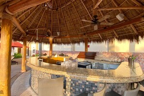 A bar area with a thatched roof, a pool, and a stone wall. - Cozy Beachfront Apartment With Stunning Ocean Views and Resort Amenities (Puerto Vallarta)