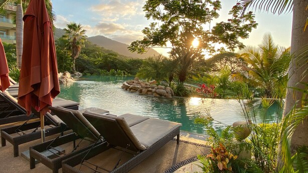 A pool area with lounge chairs and umbrellas, surrounded by lush greenery and a mountain backdrop. - Cozy Beachfront Apartment With Stunning Ocean Views and Resort Amenities (Puerto Vallarta)