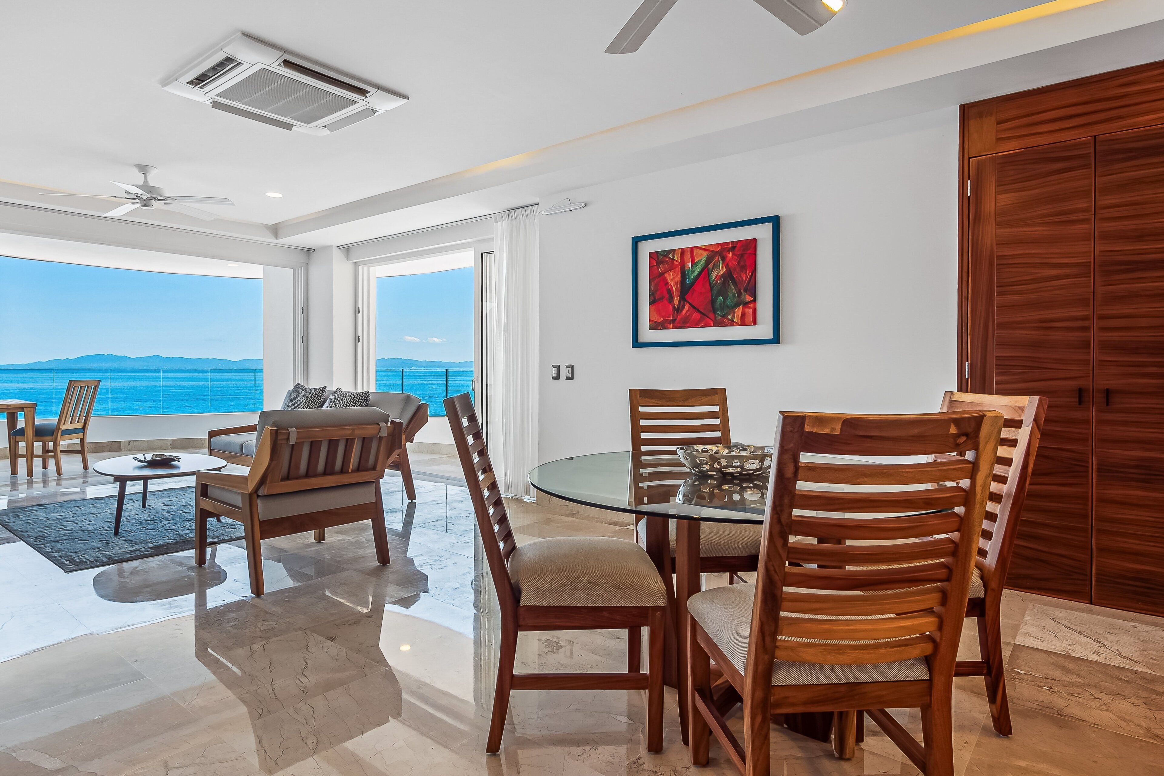 A modern dining area with a glass table, wooden chairs, and a view of the sea.