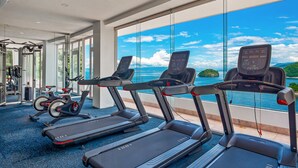 Ein Fitnessraum mit Laufbändern, Ergometer und Krafttrainingsgeräten, mit einem großen Fenster und Meerblick.