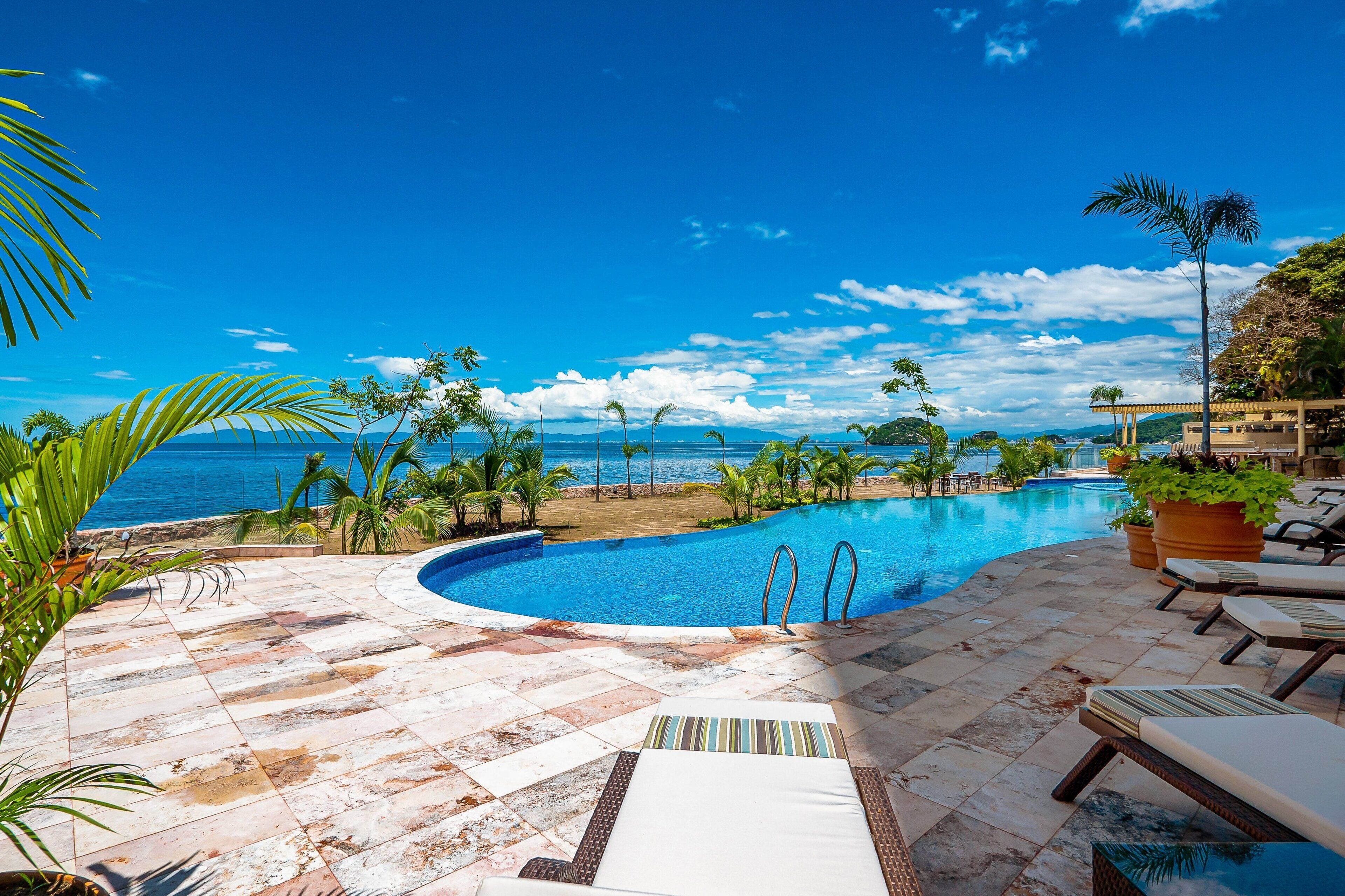 Ein Pool mit Blick auf den Ozean, umgeben von Palmen und Liegestühlen.