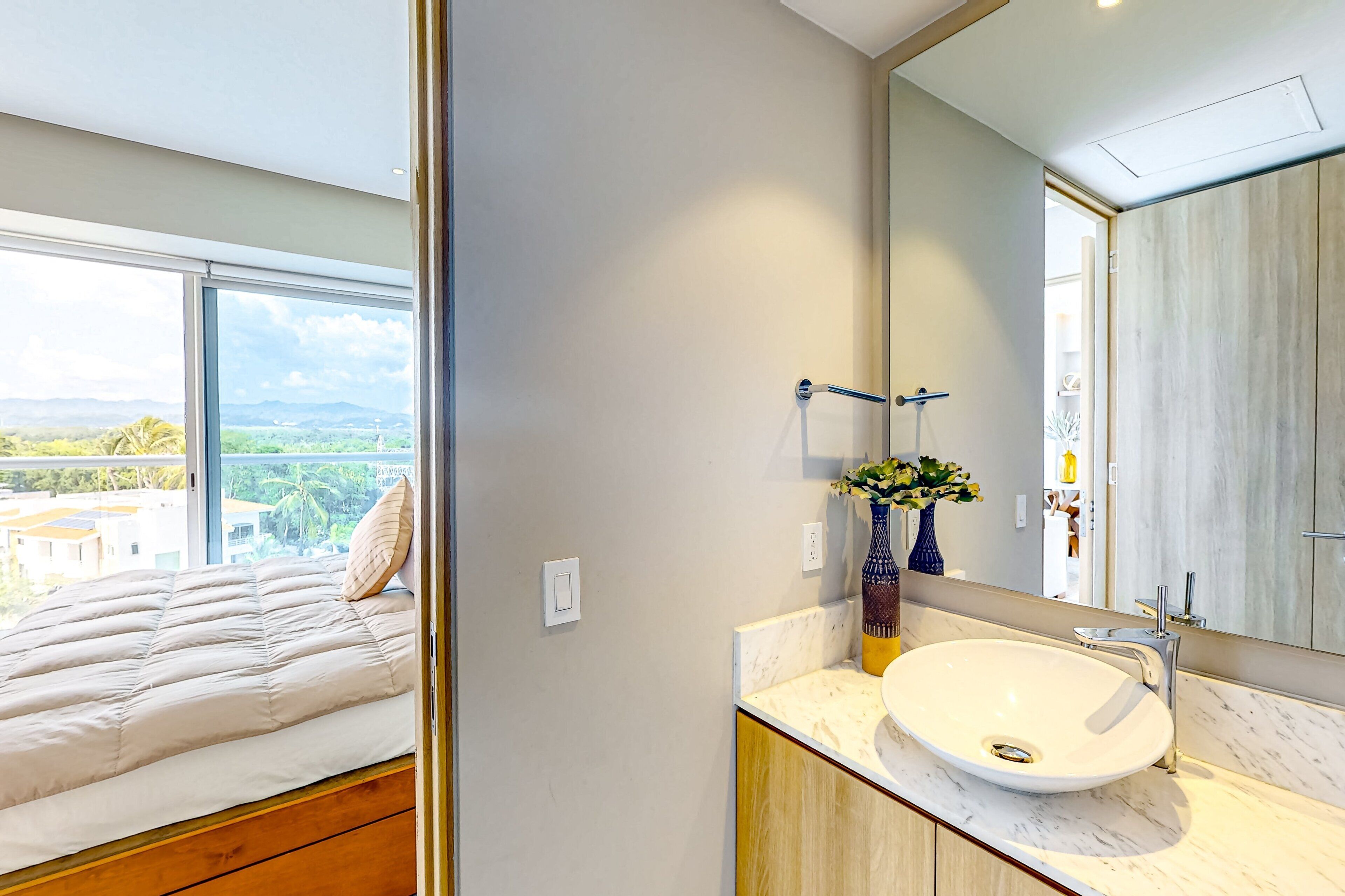 A bedroom with a bed, a bathroom with a sink and mirror, and a view of the outdoors.