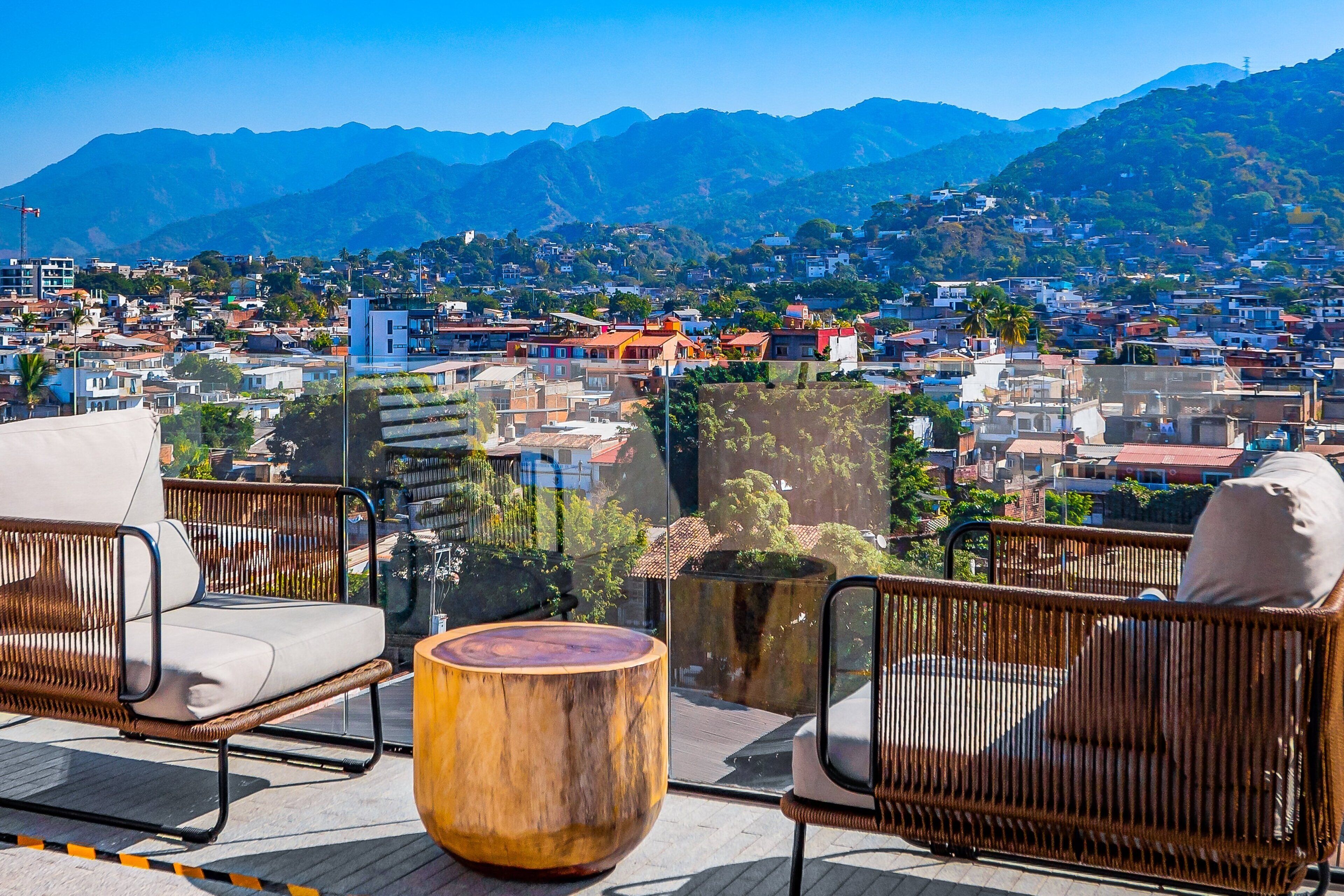 Ein Balkon mit Korbmöbeln, einem hölzernen Hocker und Blick auf die Stadt mit Bergen im Hintergrund.