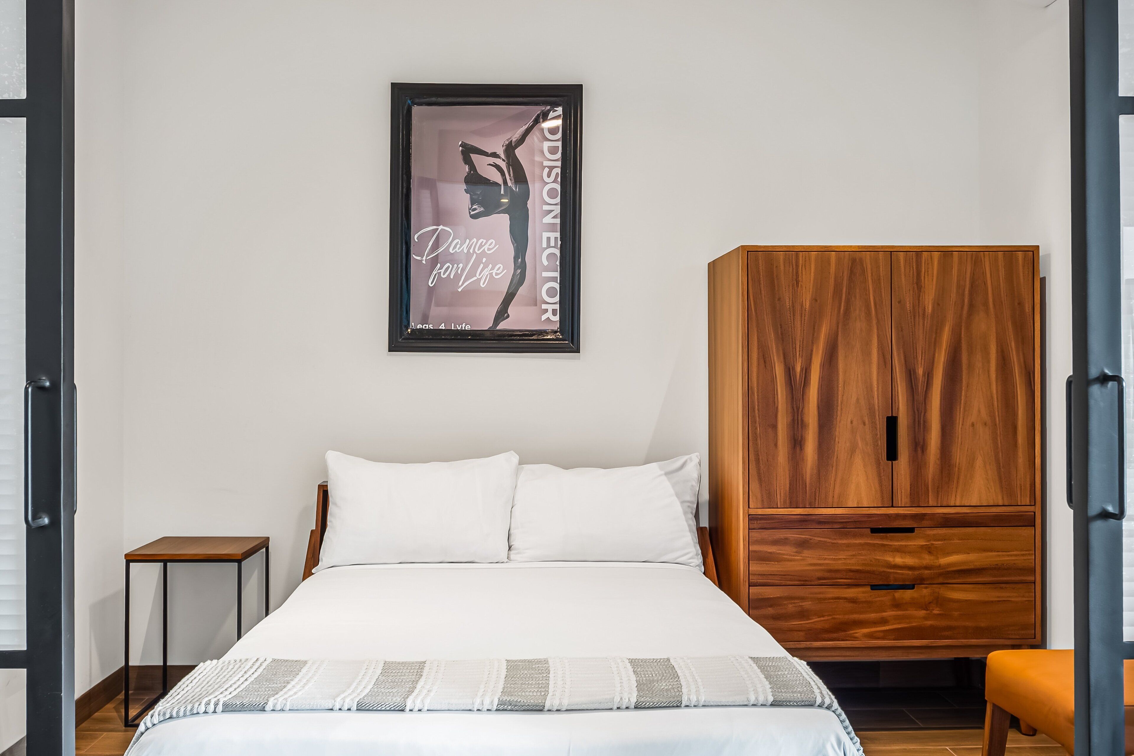 A bedroom with a bed, a wooden wardrobe, a small table, and a framed poster on the wall.
