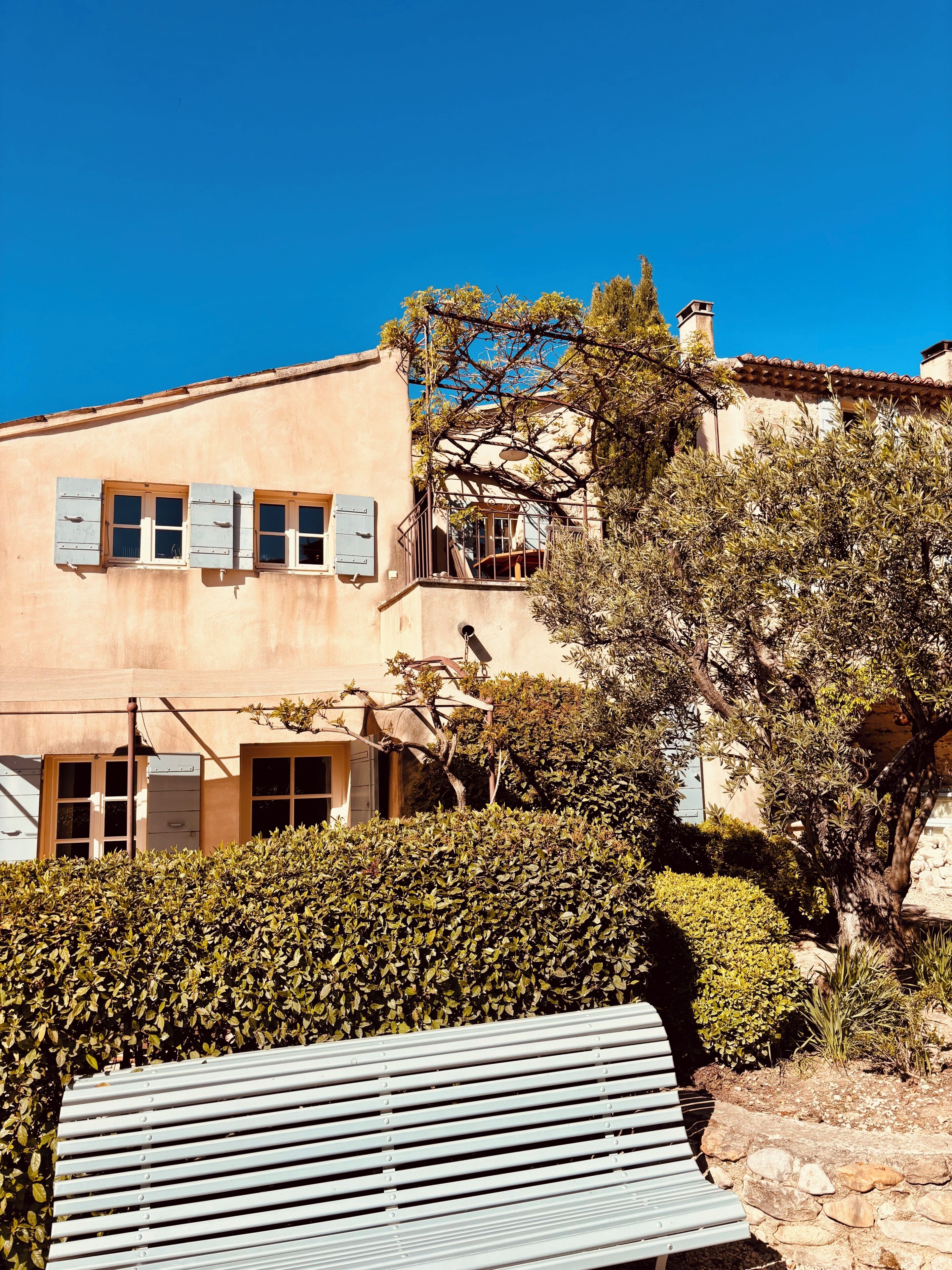 A building with a balcony, a bench, and a hedge in the foreground.