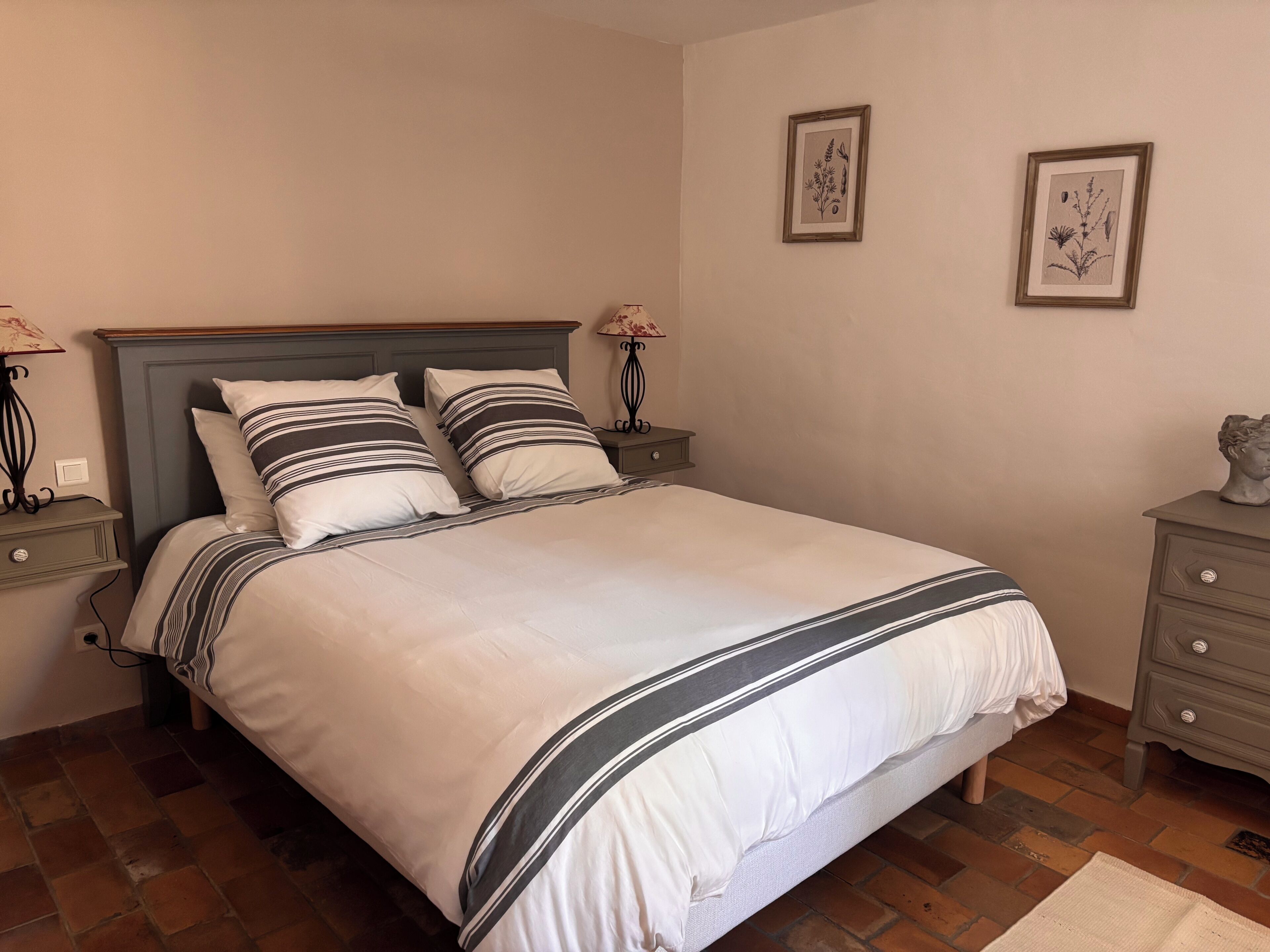 Una cama bien hecha con ropa de cama a rayas, una mesita de noche con una lámpara y un aparador con una escultura de busto.