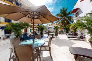 An outdoor patio area with round tables, chairs, and umbrellas, surrounded by potted plants and palm trees. - Modern & Bright Condo With Ocean Views & Pool - Walk to the Beach (Puerto Vallarta)