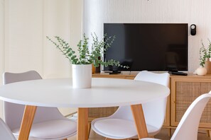 Uma sala de estar moderna com uma mesa redonda branca, cadeiras brancas, uma TV de tela plana e duas plantas em vasos.