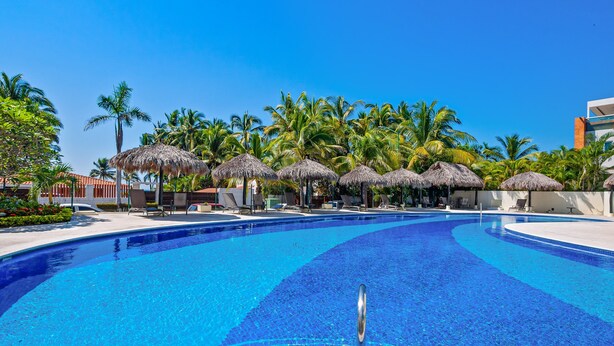 A swimming pool with a clear blue water, surrounded by palm trees and thatched umbrellas. - Stylish Condo With Covered Balcony, Pool & Pool Tables (Nuevo Nayarit)