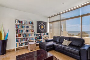 A modern living room with a leather sofa, a bookshelf, and a large window. - Stylish Condo With Covered Balcony, Pool & Pool Tables (Nuevo Nayarit)