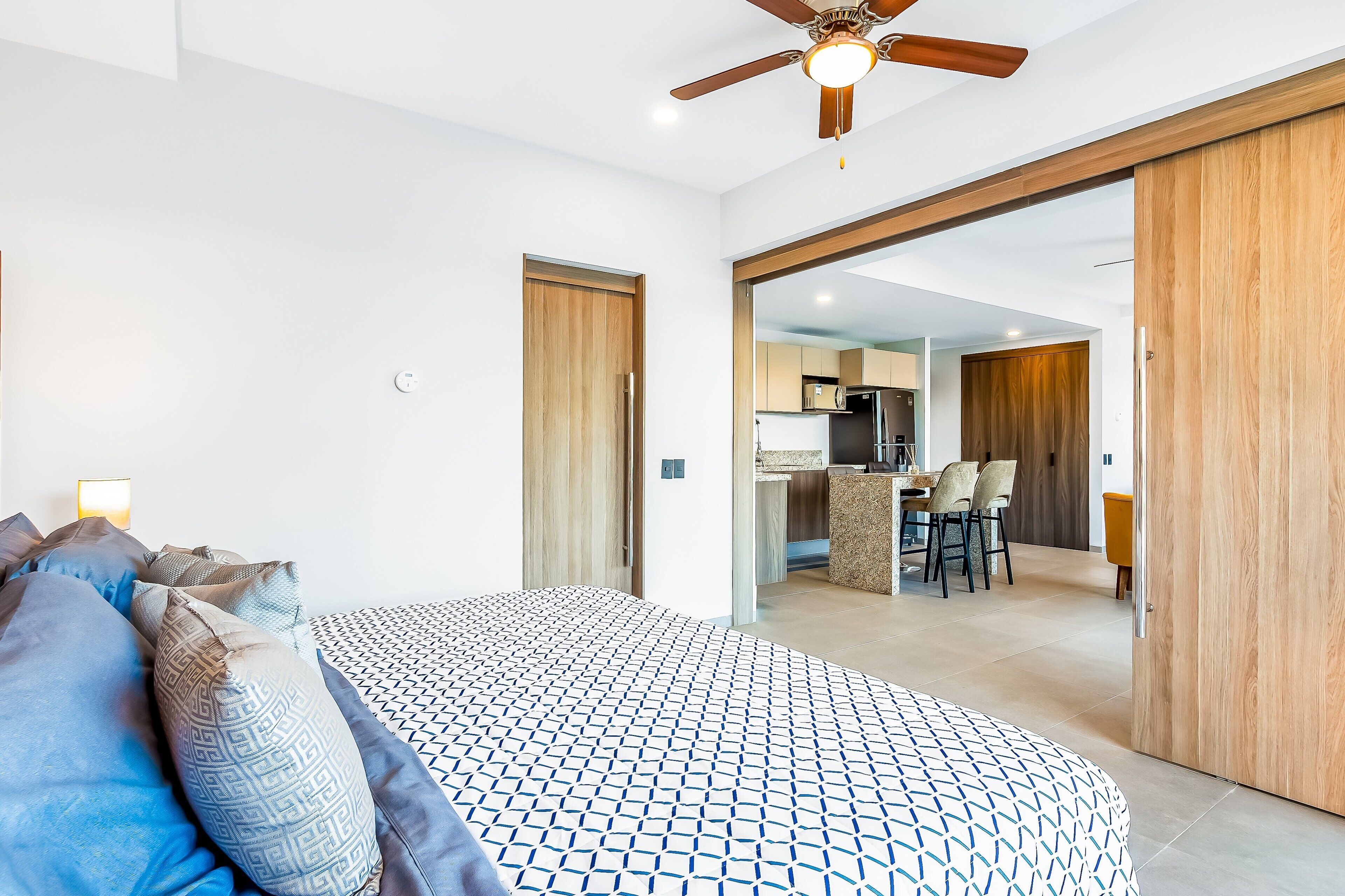 A bedroom with a bed, pillows, a ceiling fan, and a view into the kitchen area.
