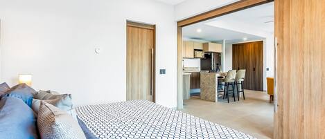 A bedroom with a bed, pillows, a ceiling fan, and a view into the kitchen area.
