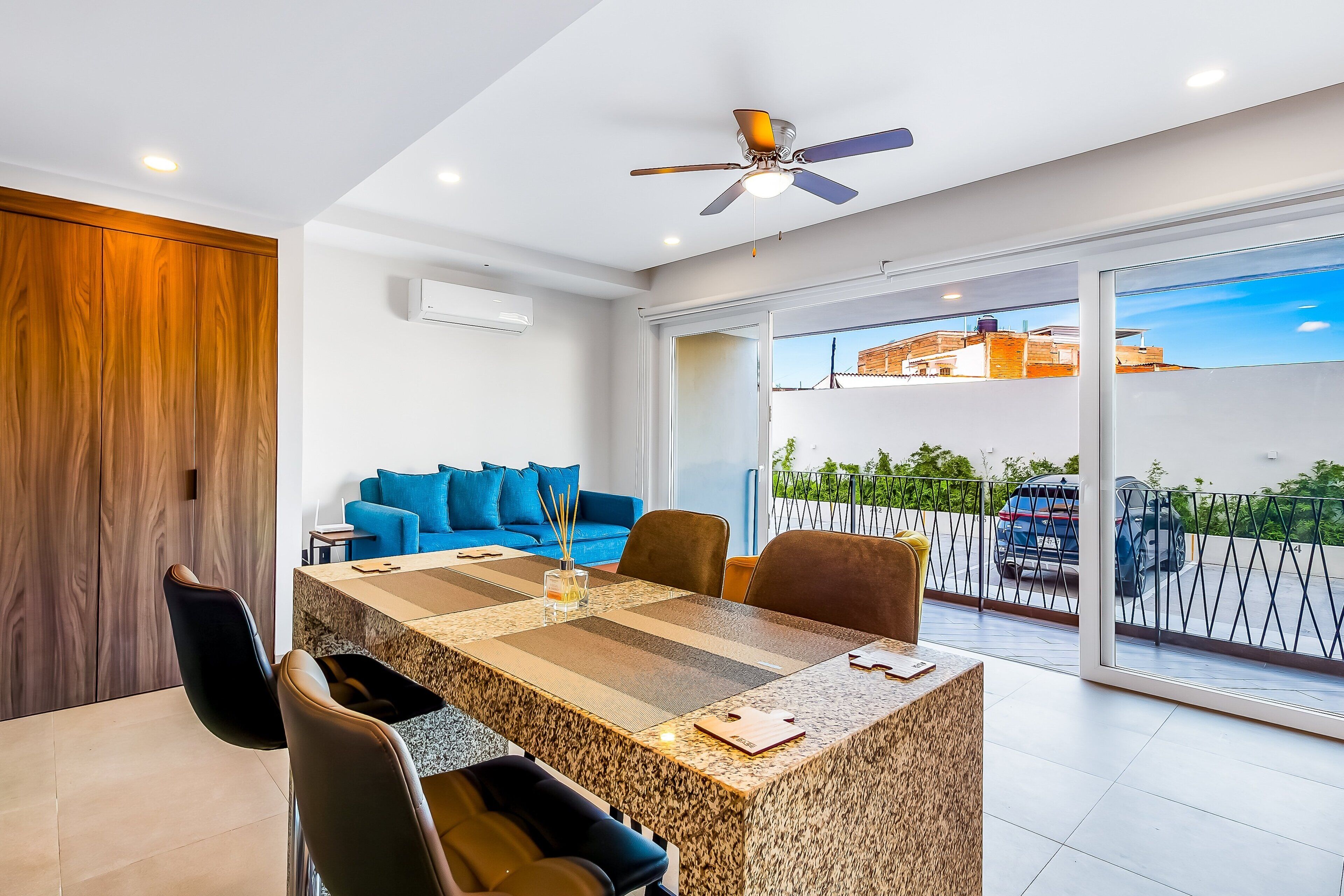 A modern living room with a large table, comfortable seating, a ceiling fan, and a balcony with a view.