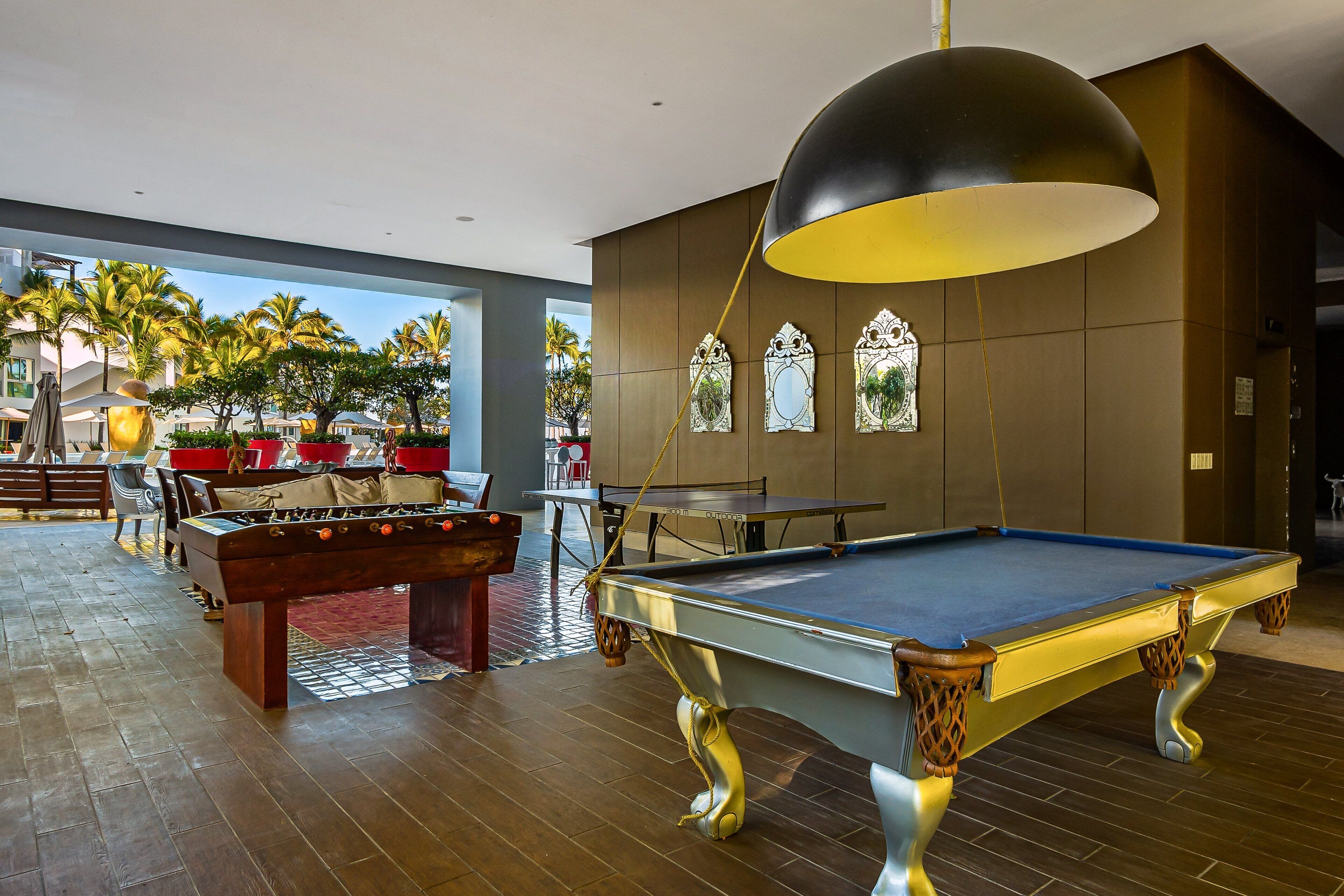 Uma mesa de bilhar com design exclusivo, uma mesa de futebol de mesa e uma luminária pendente grande em um ambiente com vista para palmeiras.