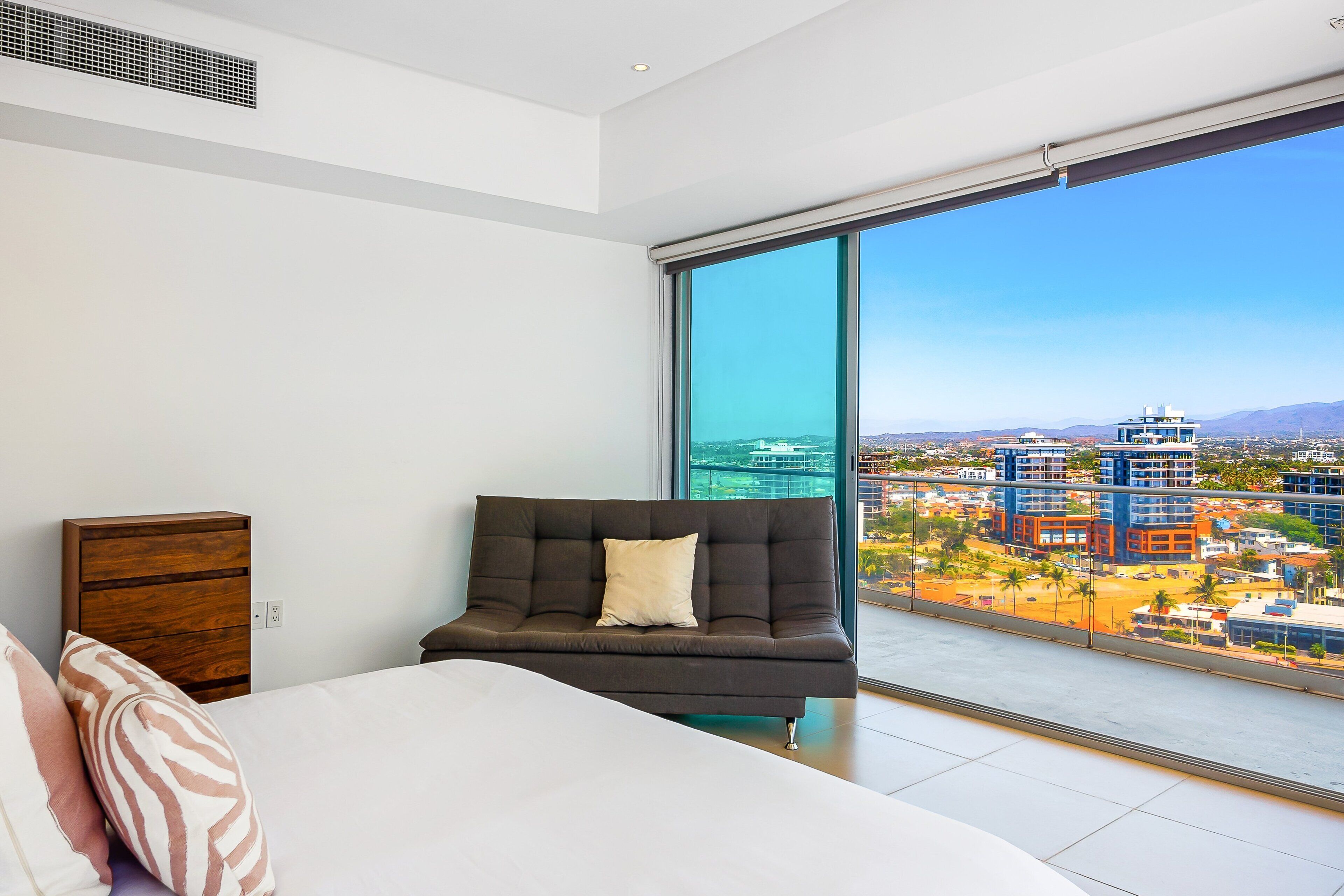 A modern bedroom with a large bed, a dark sofa, and a view of a cityscape through a large window.