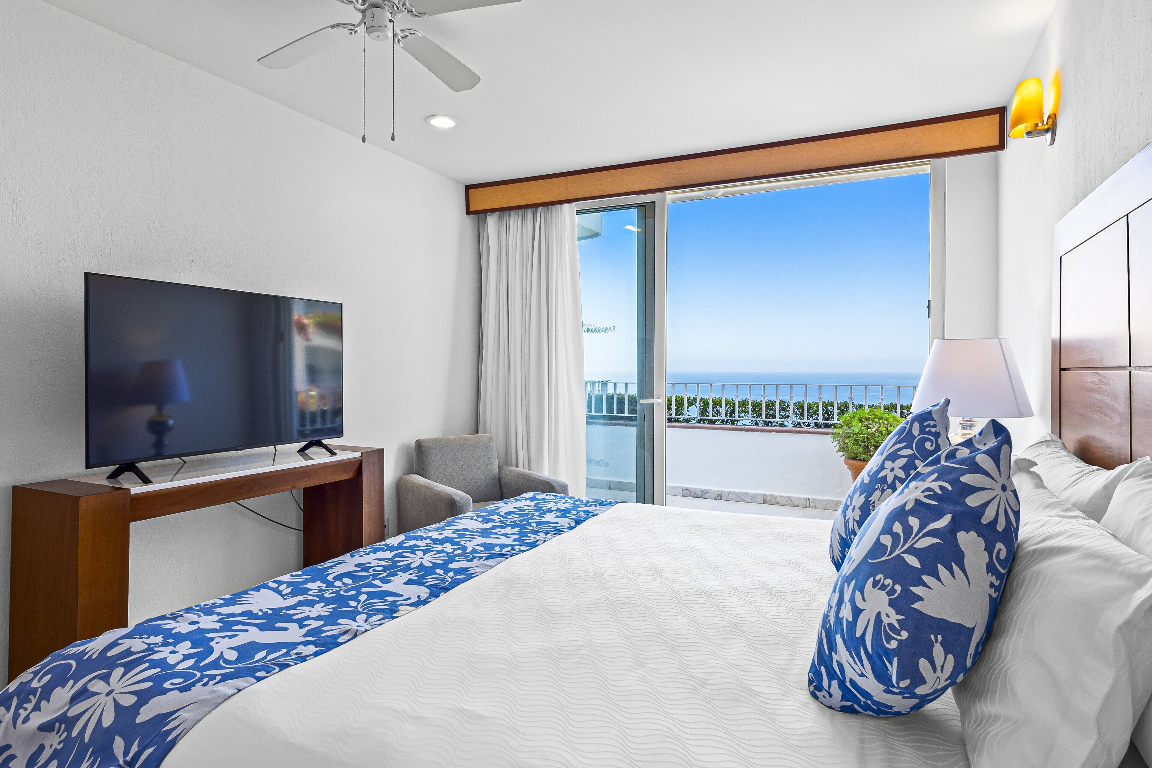 Ein Hotelzimmer mit Bett, Fernseher, Schreibtisch, Stuhl und einem Balkon mit Meerblick.