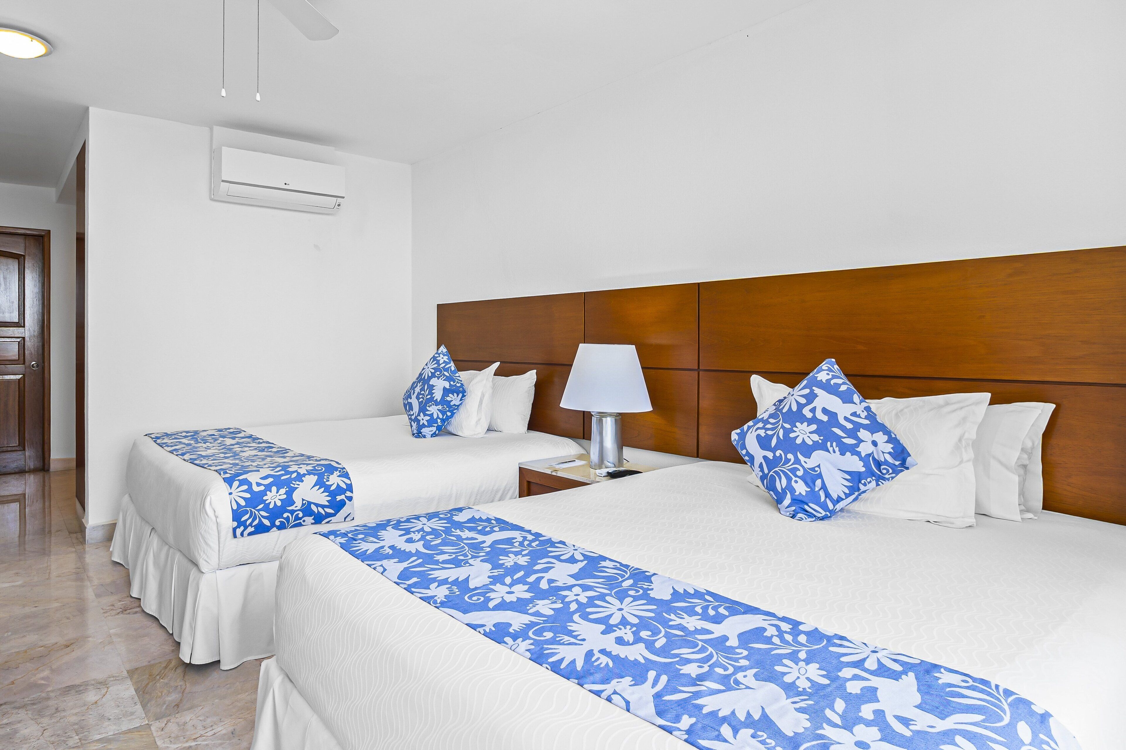 Una habitación de hotel con dos camas, cabecero de madera, mesita de noche, lámpara y equipo de aire acondicionado.