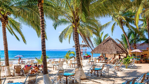 A beach with palm trees, lounge chairs, and a thatched-roof structure. - Stylish Beach Condo With Balcony View, Pool, Wifi & Central AC (Nuevo Nayarit)