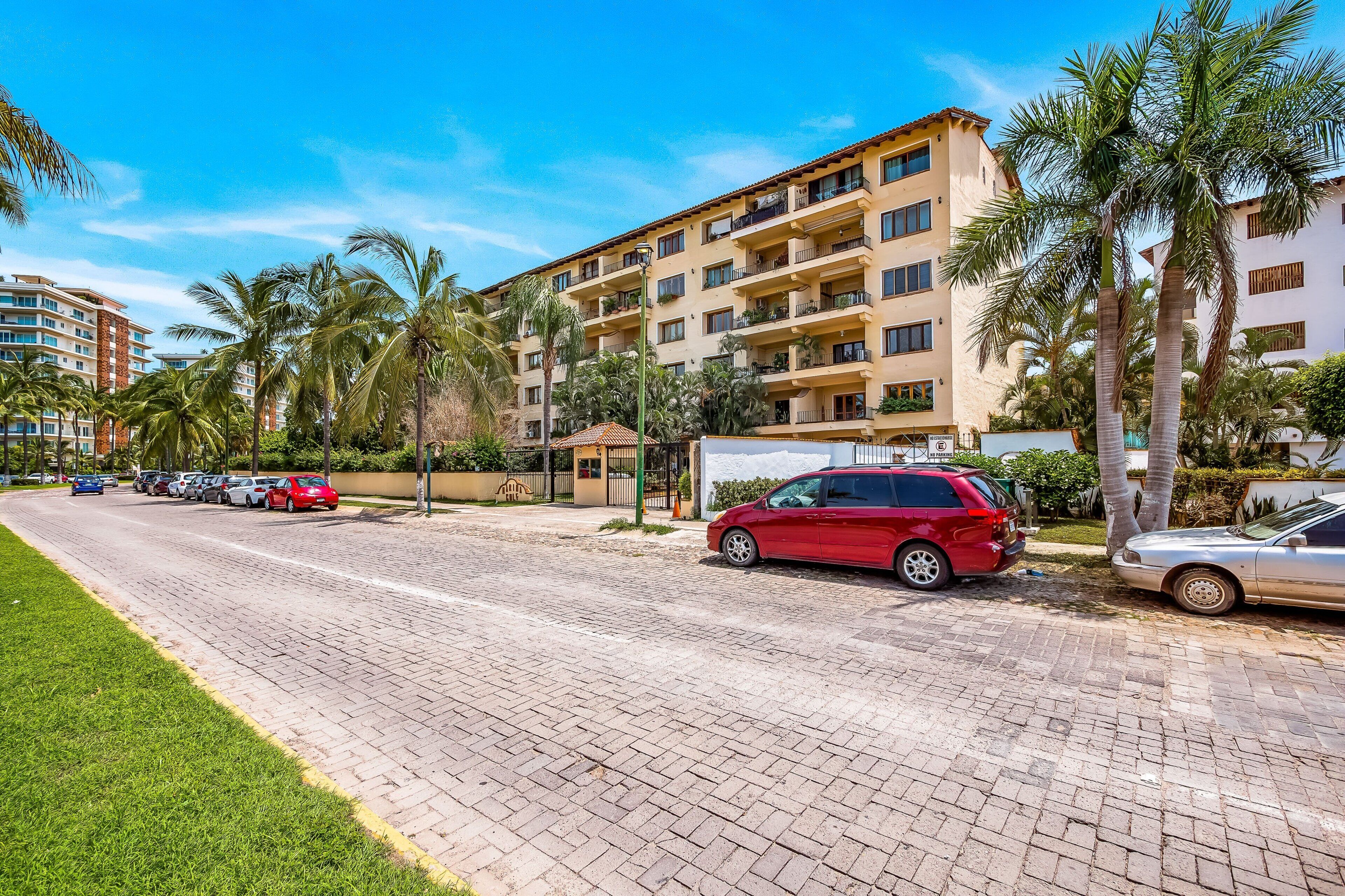 A multi-story hotel with a paved driveway, parked cars, and palm trees.