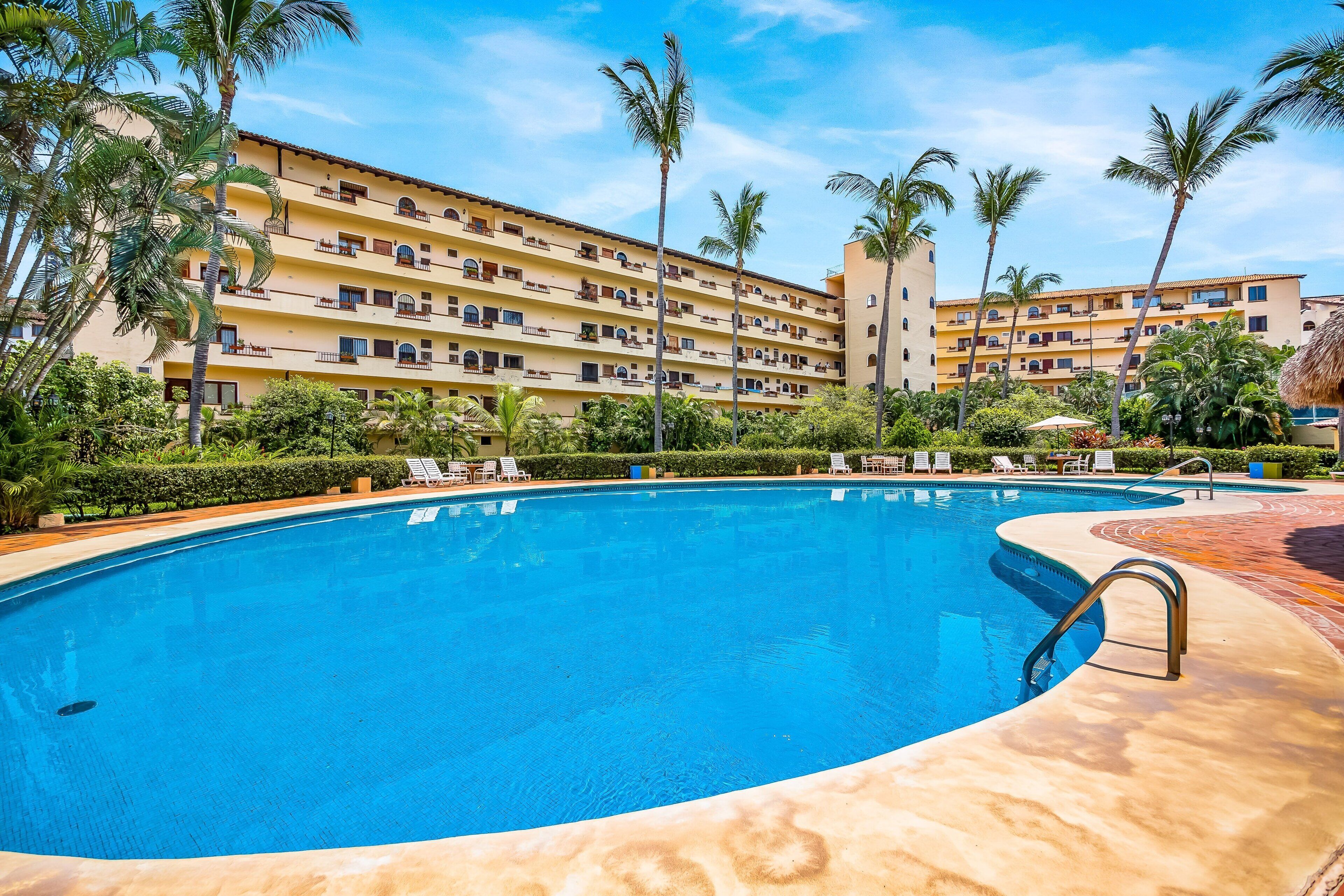 A hotel with a swimming pool, palm trees, and a clear blue sky.