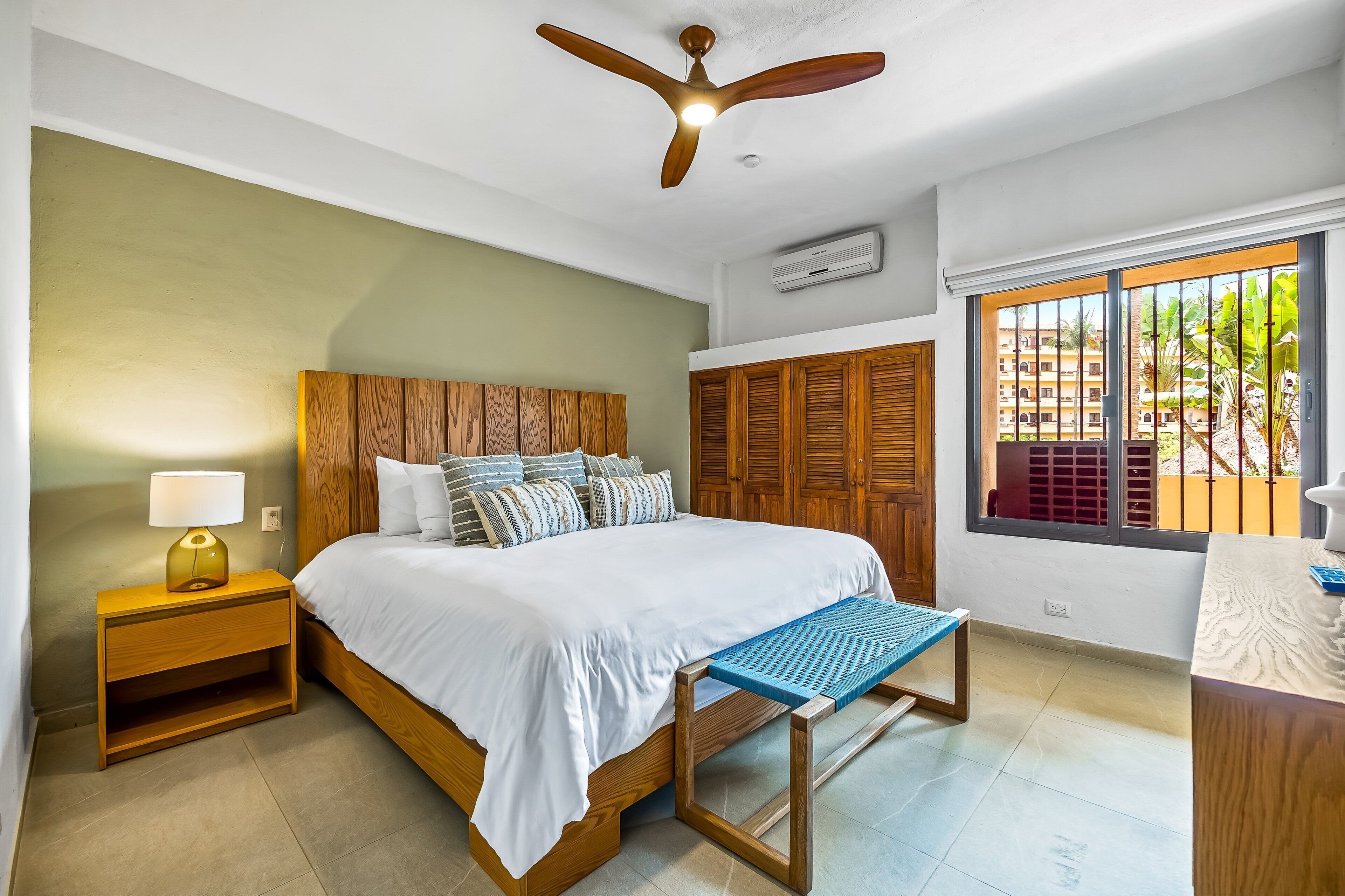 Une chambre à coucher comprenant un lit, une tête de lit en bois, une table de chevet avec une lampe, une fenêtre avec vue, un ventilateur de plafond et une armoire en bois.