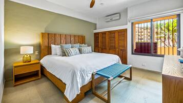 Une chambre à coucher comprenant un lit, une tête de lit en bois, une table de chevet avec une lampe, une fenêtre avec vue, un ventilateur de plafond et une armoire en bois.