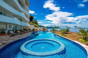 Piscina com padrão de mosaico circular, rodeada por espreguiçadeiras e plantas em vasos, com vista para uma montanha e o mar.