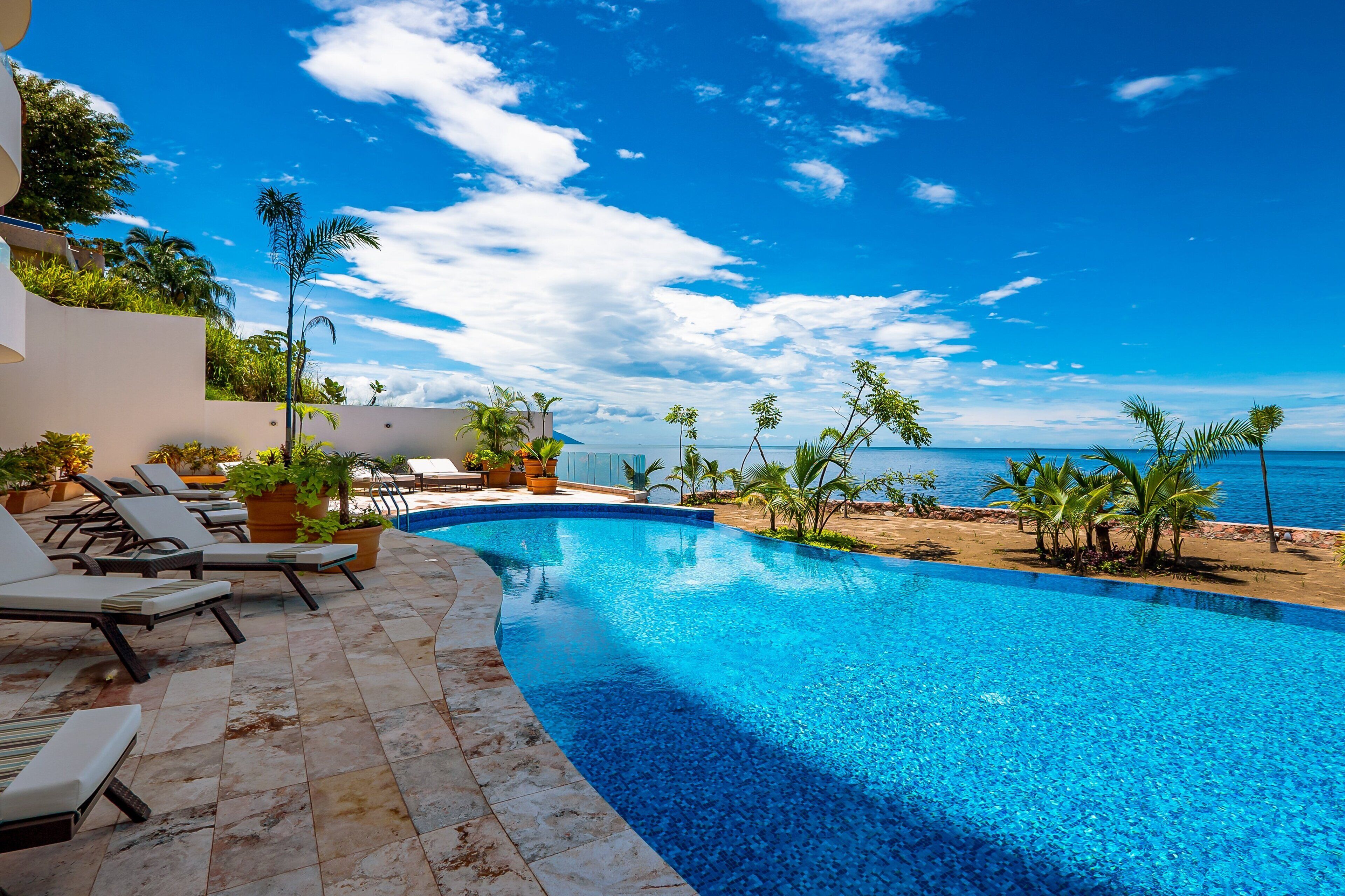 A curved pool with lounge chairs and potted plants, overlooking a beach and ocean.