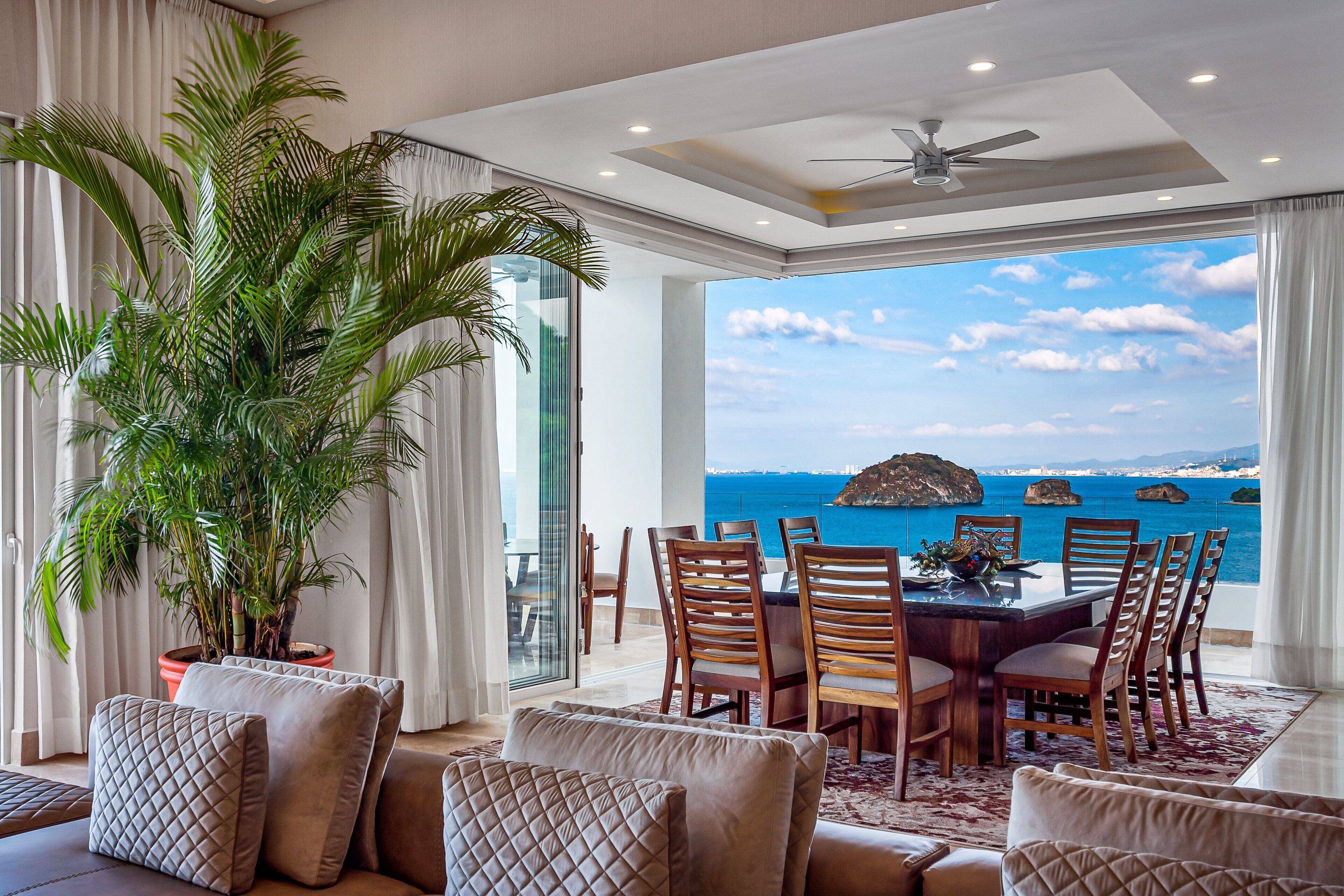 A modern dining area with a view of the sea, a large potted plant, and a ceiling fan.