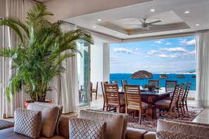 A modern dining area with a view of the sea, a large potted plant, and a ceiling fan. - Upscale Oceanfront Condo With Extensive Balcony, Pool & Ocean Views (Puerto Vallarta)