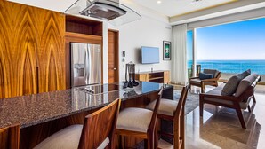 A modern kitchen with a granite countertop, wooden cabinetry, and a built-in oven. There is a dining area with a bar and seating, and a large window offering a view of the ocean. - Luxury Bayfront Condo With Amazing Views, Pool & spa (Puerto Vallarta)