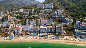 A coastal cityscape with high-rise hotels, a beach with umbrellas, and lush green hills in the background. - Stylish Coastal Condo With Pool, Private Balcony & Stunning Ocean Views (Puerto Vallarta)