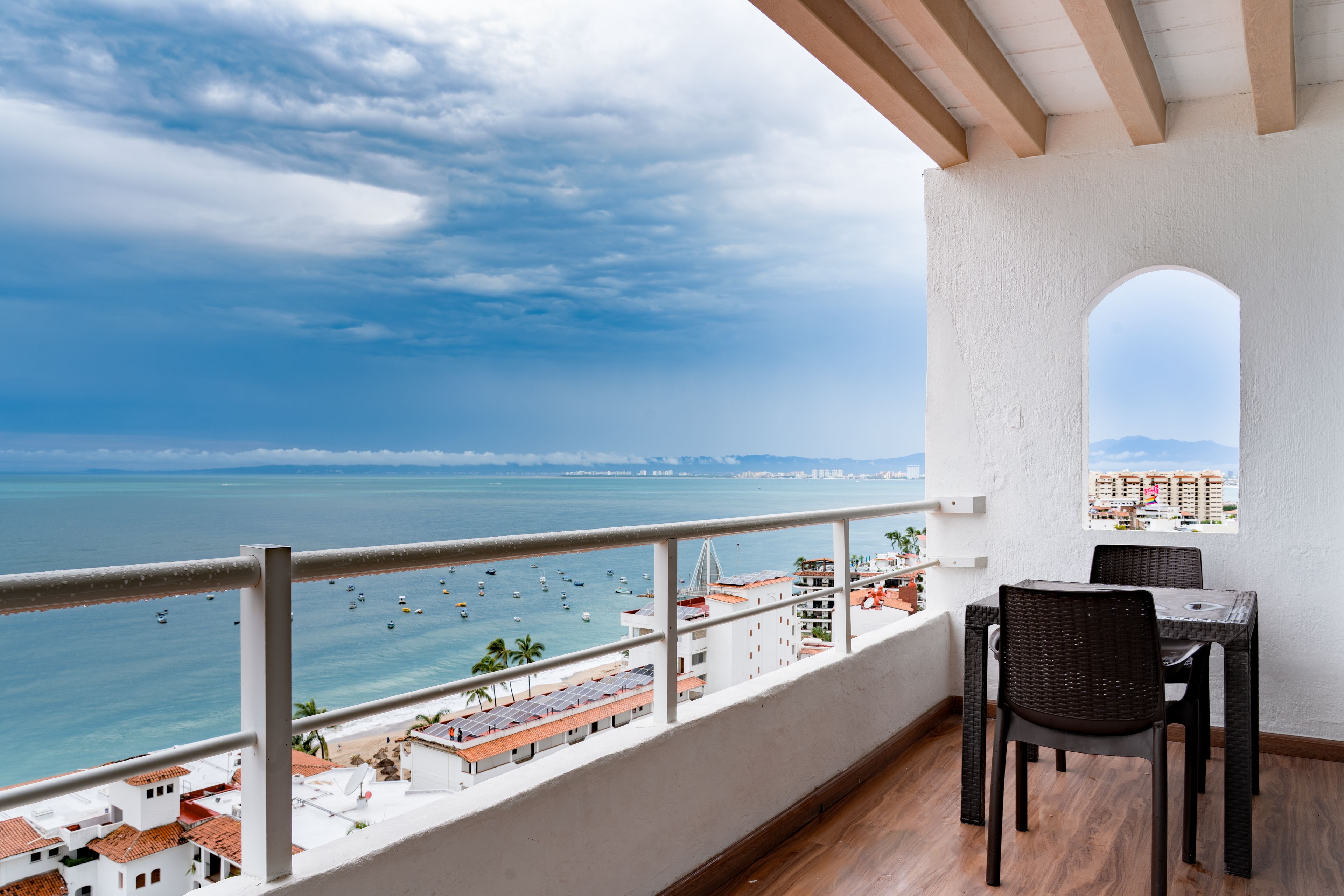 Un balcón con vista al mar, una mesa con sillas y una pared blanca con una ventana.