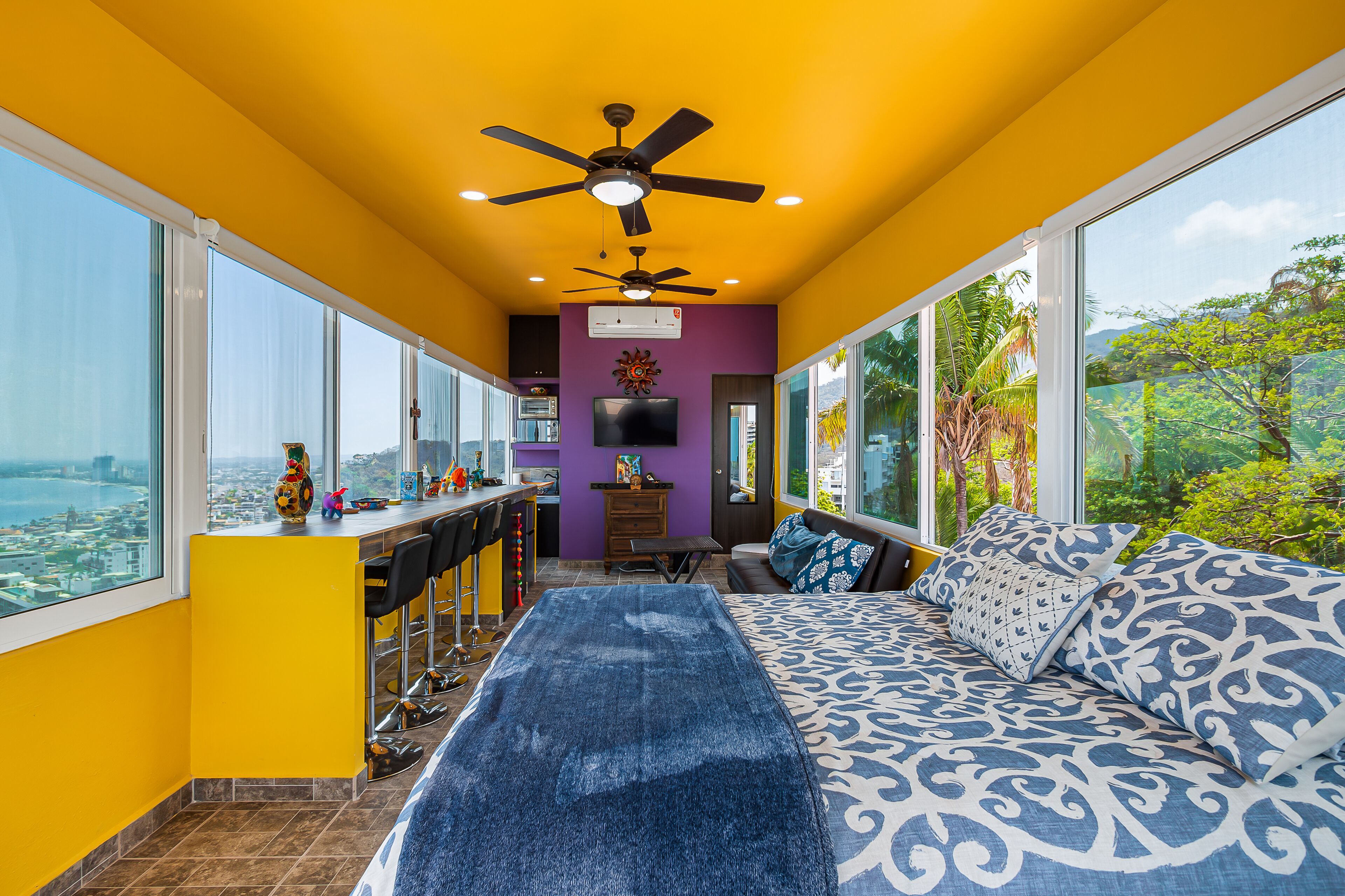 A room with a bed, a bar area, and a large window with a city view.
