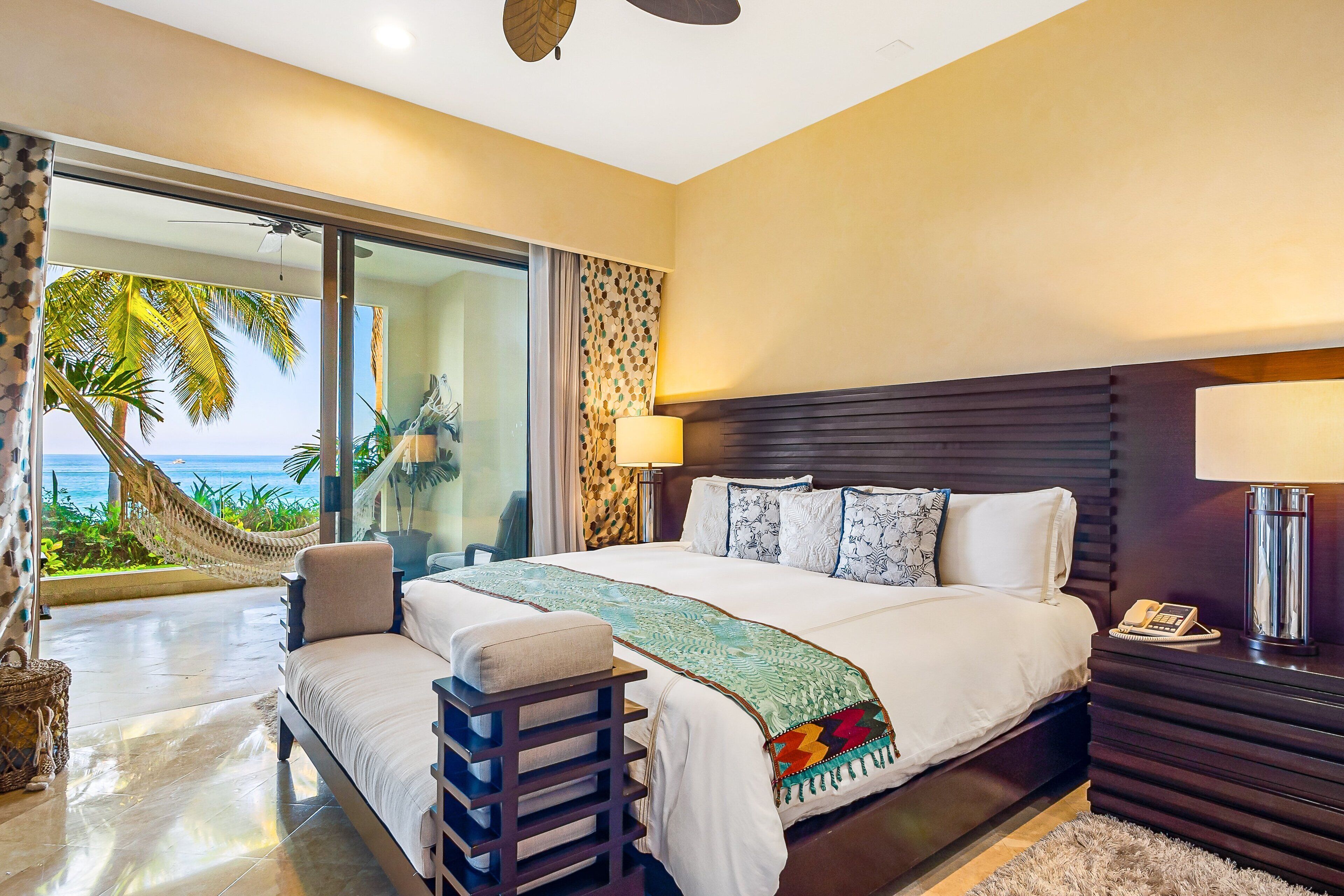 A hotel room with a bed, a nightstand, a lamp, a view of the ocean, and a hammock.