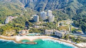 Un complejo turístico frente a la playa con villas, un hotel central y montañas al fondo.